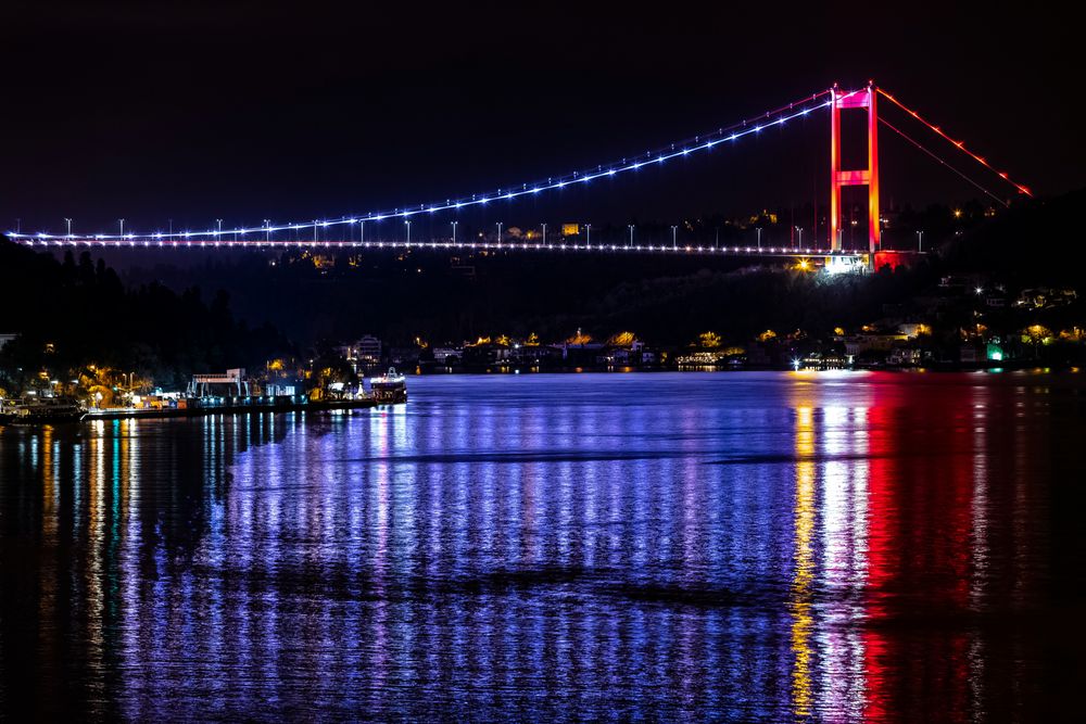 Die beleuchtete Fatih-Sultan-Mehmet-Brücke über dem Bosporus bei Nacht, gespiegelt im Wasser